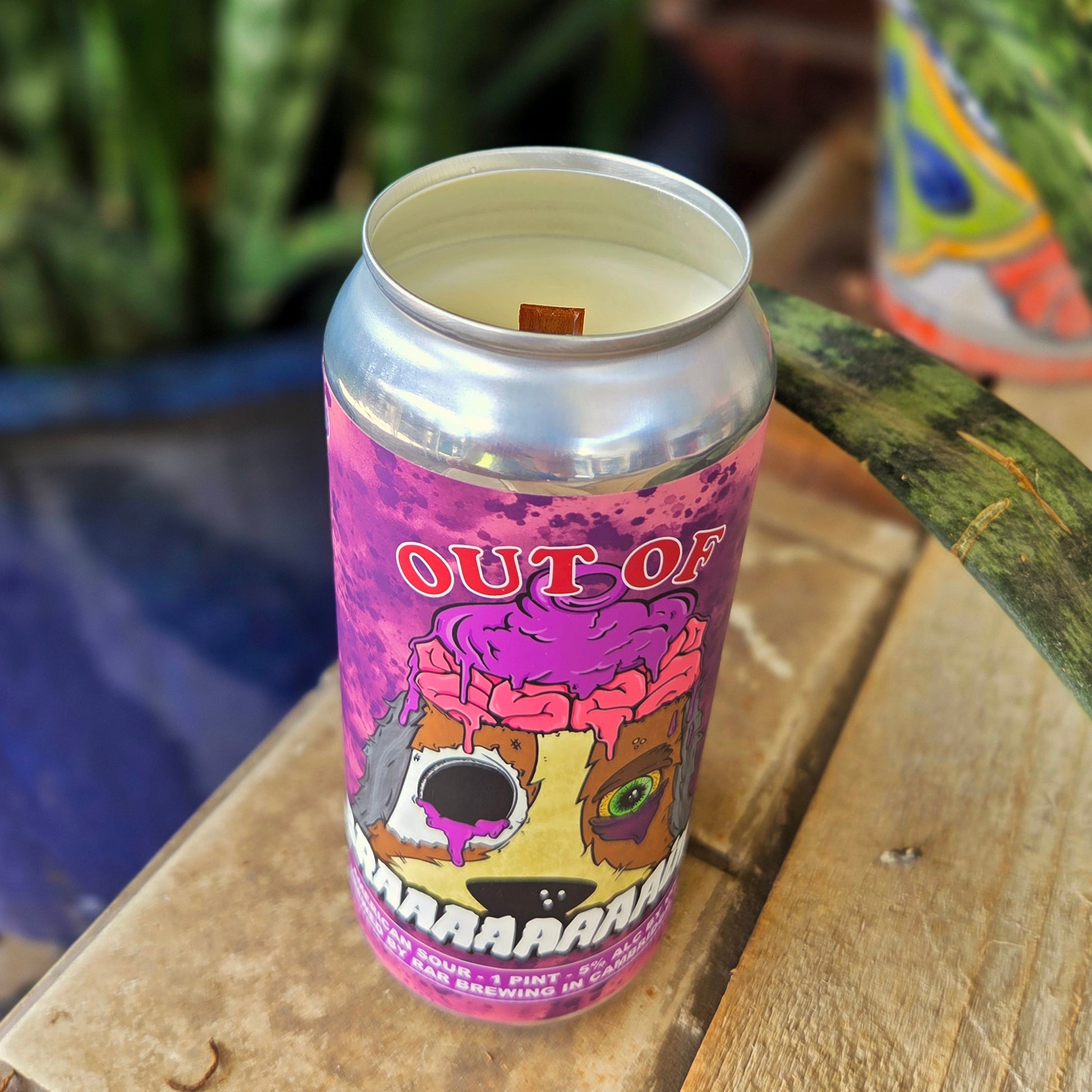 Angled shot of a beer can candle with a vivid purple “Out of BRAAAAAAAAins” label featuring a cartoon dog with an exposed pink brain, torn eye, and oozing details. This unique upcycled candle is hand-poured using coconut soy wax and a crackling wooden wick.