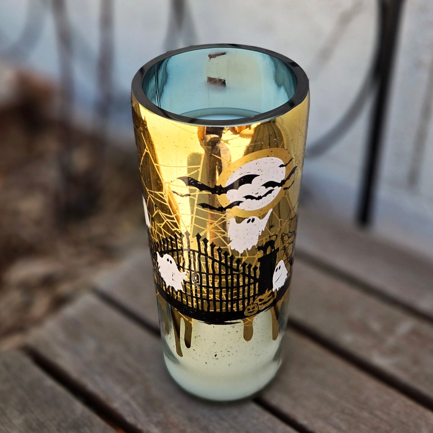 Angled shot of a Halloween-themed wine bottle candle with a golden exterior featuring spooky artwork of ghosts, bats, and a haunted gate. This upcycled vessel is hand-poured with coconut soy wax and a wooden wick.