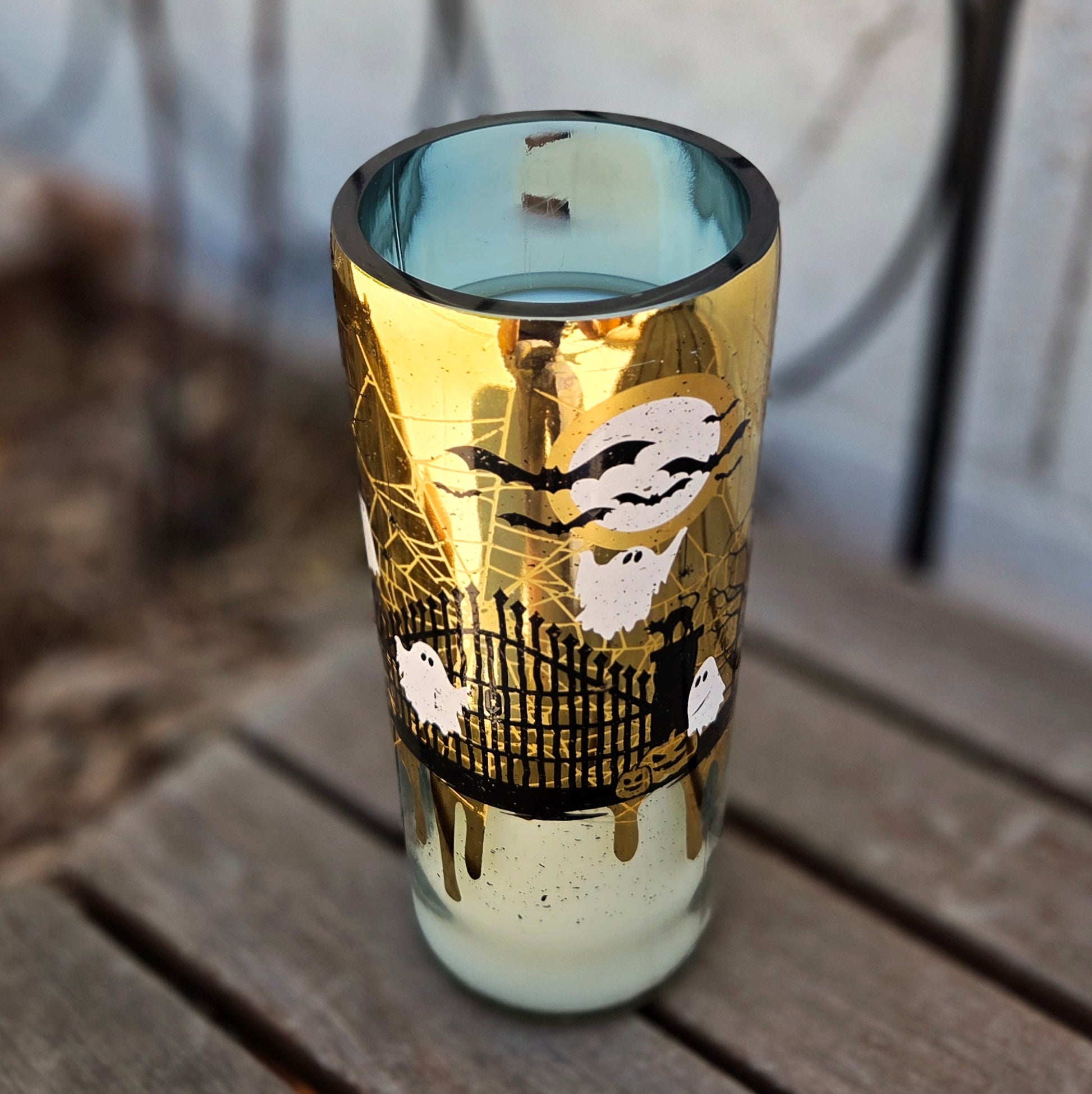 Angled shot of a Halloween-themed wine bottle candle with a golden exterior featuring spooky artwork of ghosts, bats, and a haunted gate. This upcycled vessel is hand-poured with coconut soy wax and a wooden wick.