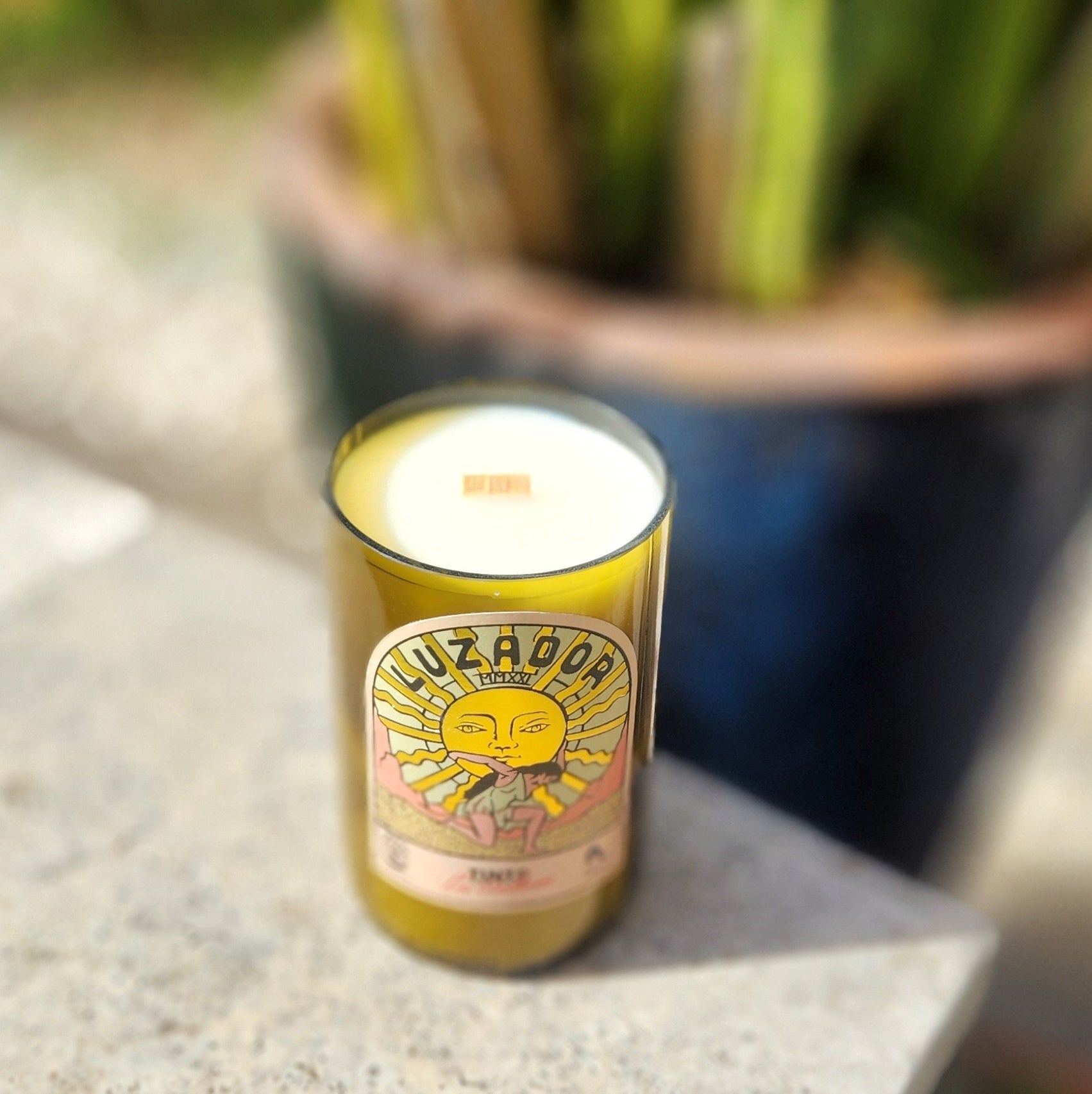 Angled view of a hand-poured coconut soy candle housed in a recycled green wine bottle with a smooth glass rim. The label features a stylized woman beneath a radiant yellow sun with bold retro typography that reads “Luzador.” A wooden wick peeks from the creamy wax, highlighting the candle’s eco-friendly and artisan-crafted charm.