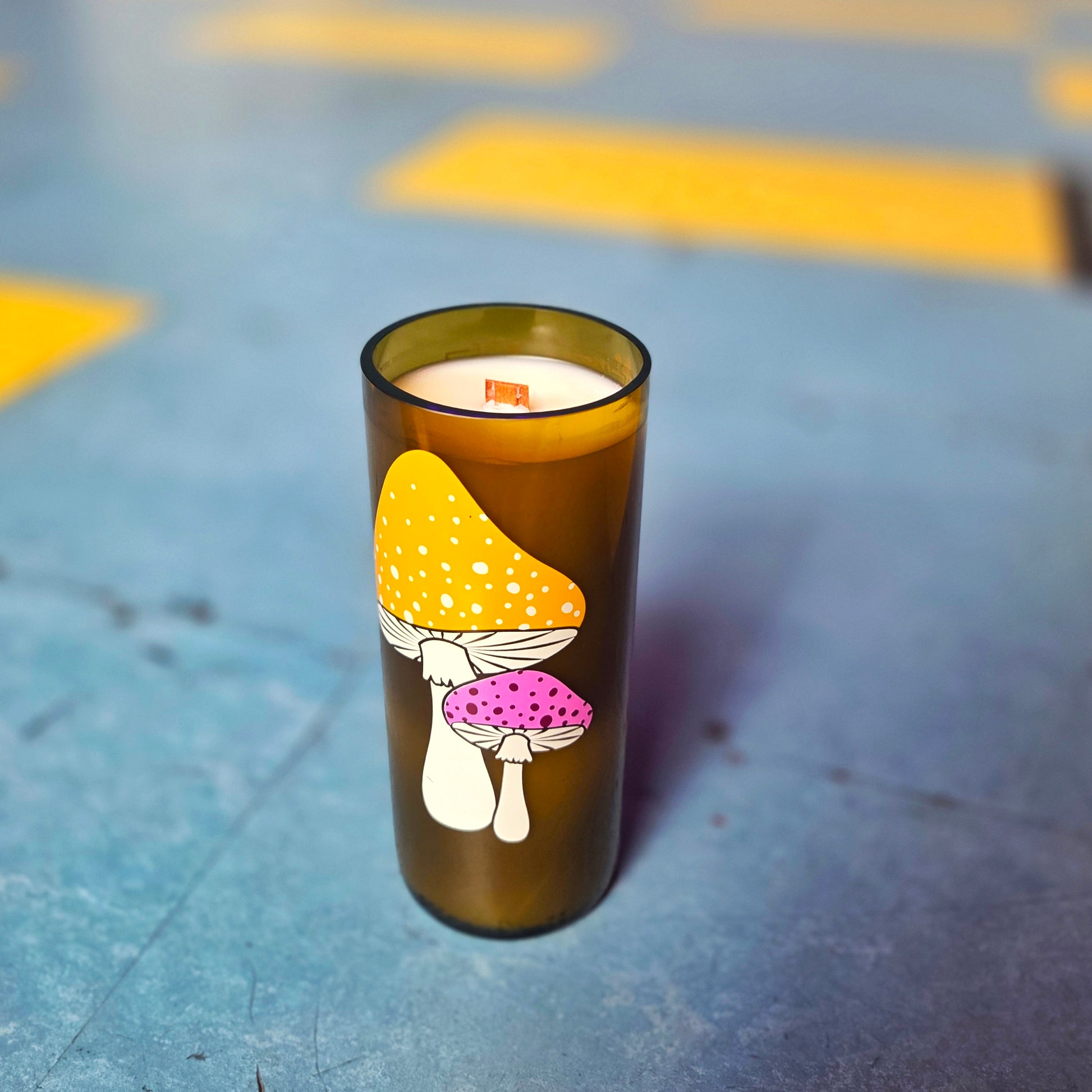 Eco-friendly wine bottle candle featuring an illustration of yellow and pink spotted mushrooms. The candle is hand-poured with creamy coconut soy wax and includes a wooden wick for a clean burn. Set against a blue and yellow background for a playful vibe.