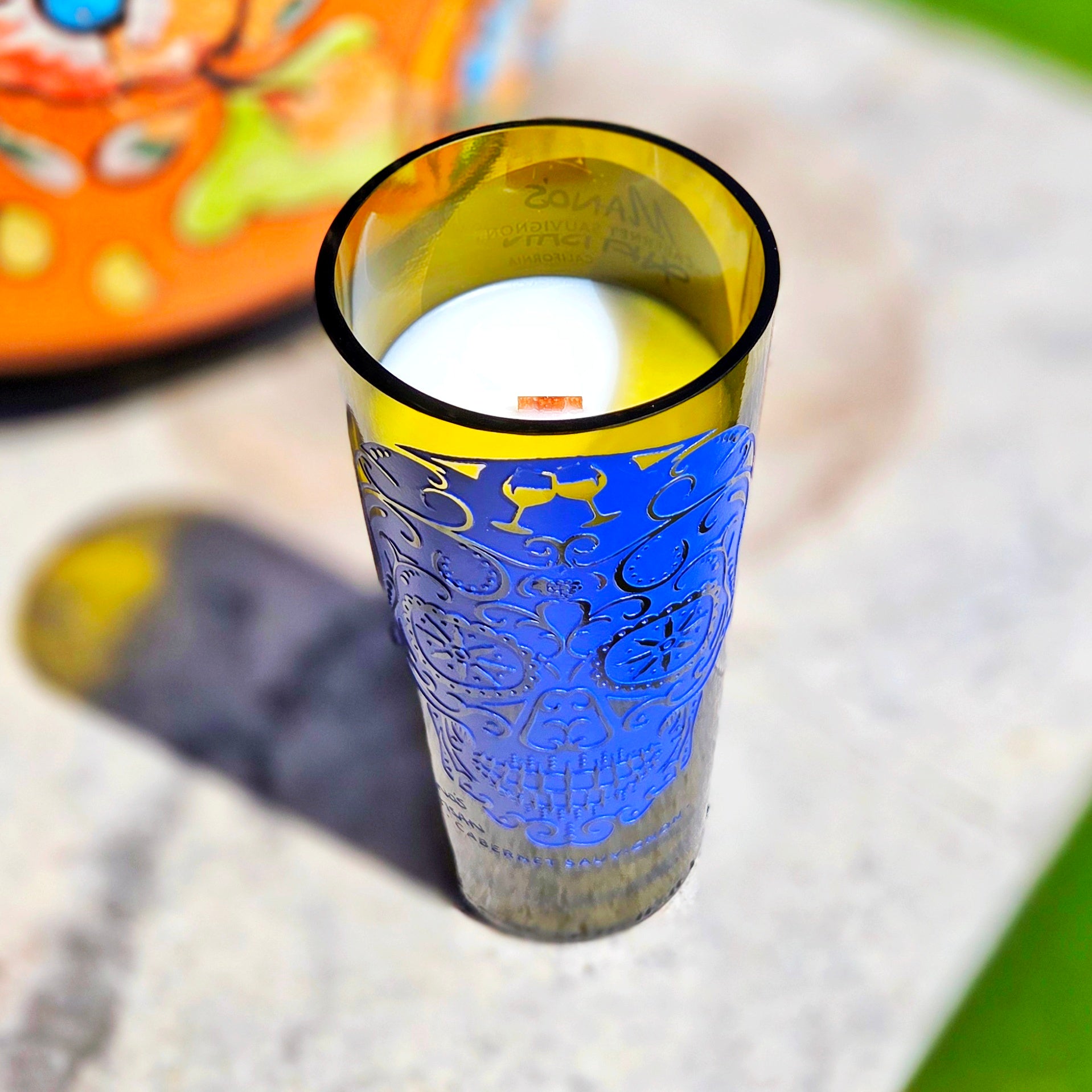Angled shot of a tall wine bottle candle featuring a blue sugar skull design, showcasing the creamy white coconut soy wax and centered wooden wick inside a smooth-cut, upcycled bottle.