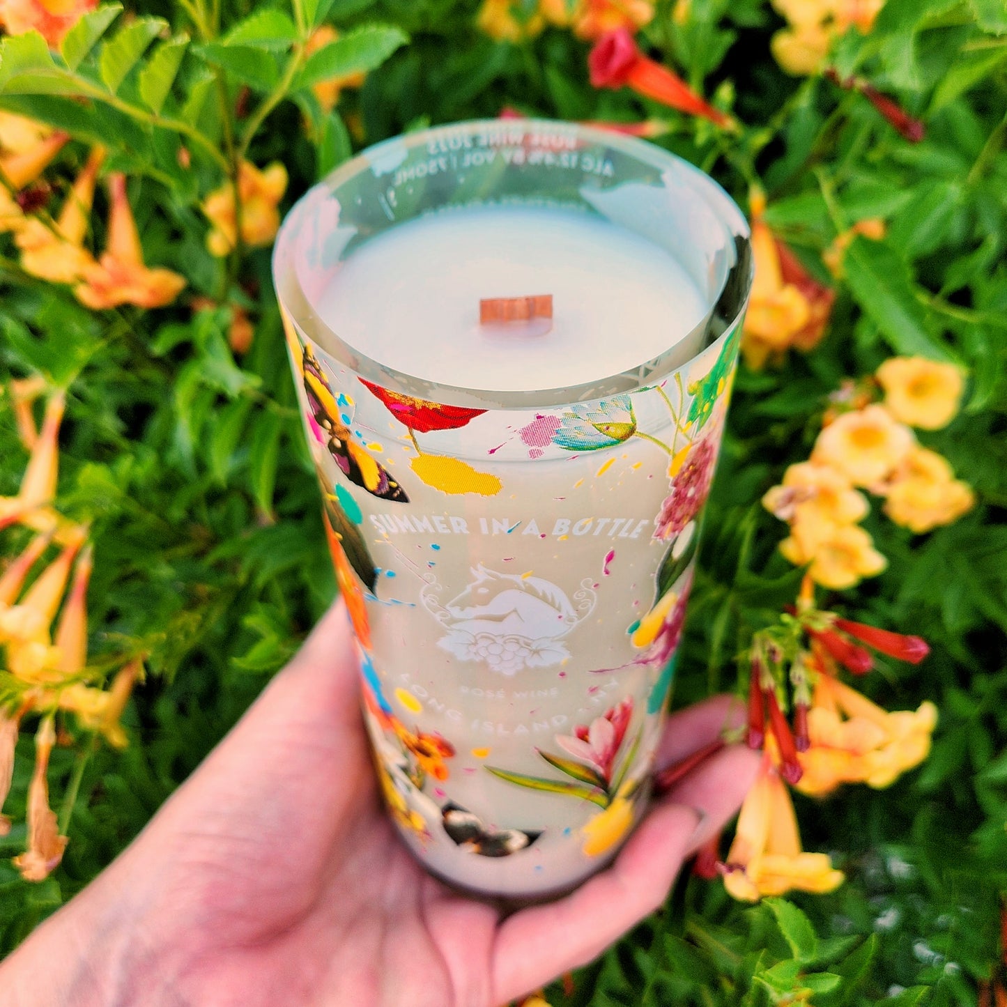 A clean-burning wooden wick candle in a recycled rosé wine bottle, topped with creamy coconut soy wax. The glass features bright floral and butterfly motifs with the words “Summer in a Bottle,” showcasing its artisan craftsmanship and sustainable design.
