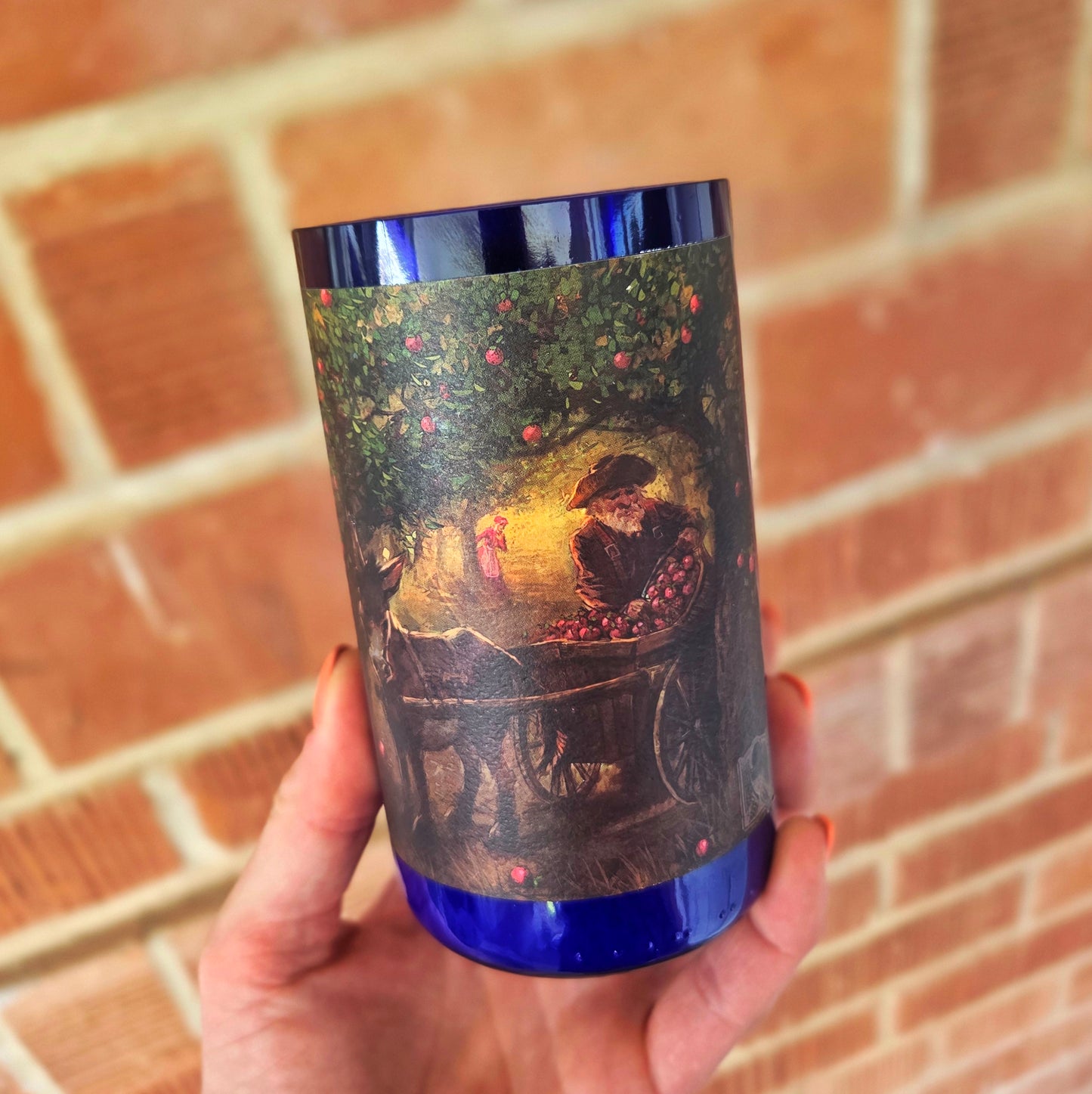 Front-facing image of a wine bottle candle wrapped in a rustic apple orchard scene, crafted in a deep blue recycled bottle and hand-poured with coconut soy wax and a wooden wick.