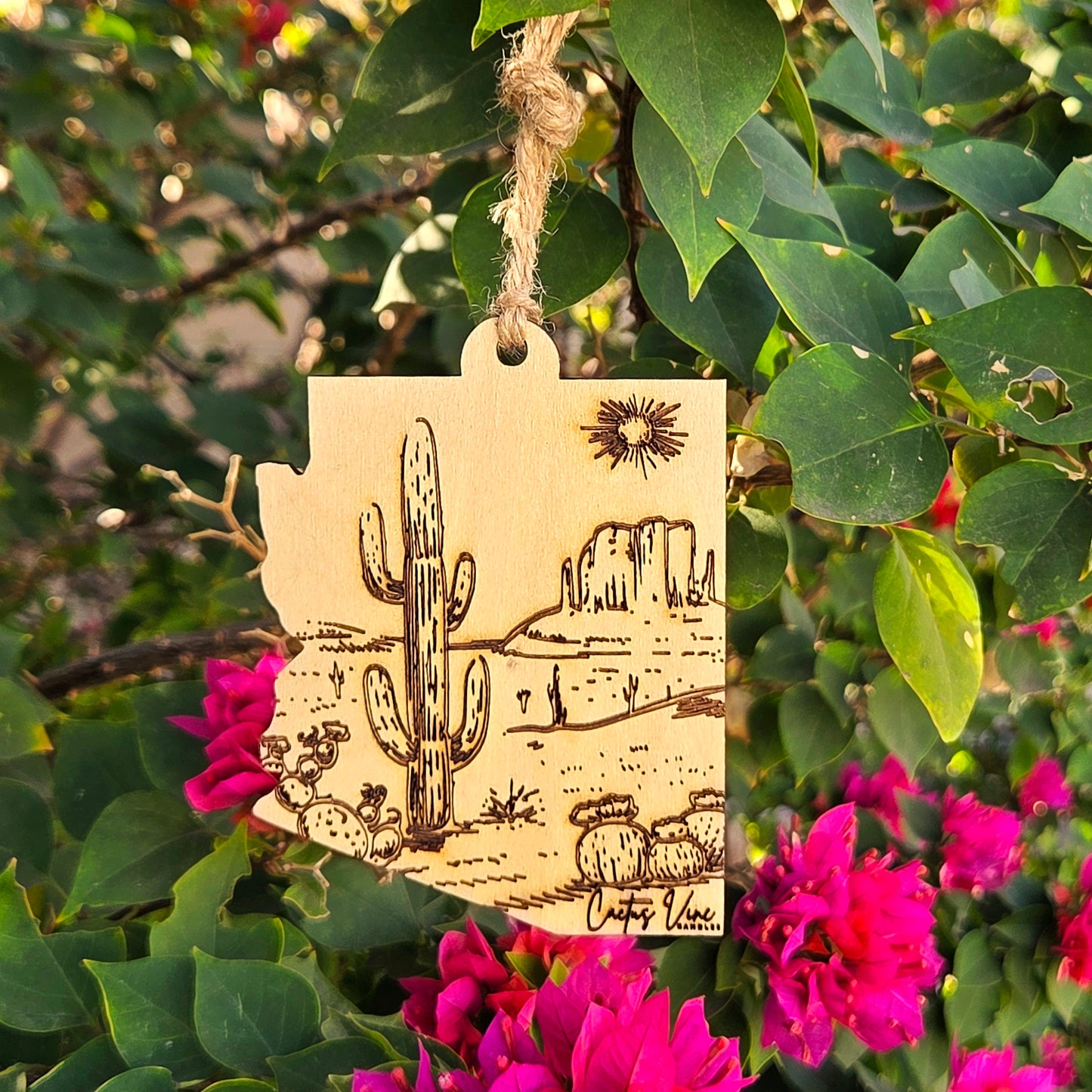 Wooden, reusable air freshener in the shape of Arizona with a cactus and desert etching.  Hanging on lush green and pink plants.