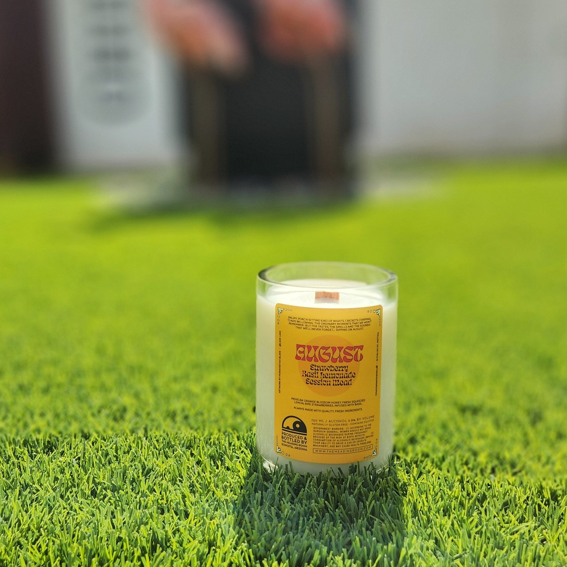 The back of the August candle featuring the original mead label that reads “Elderberry Basil Lemonade Session Mead.” The candle sits on a green grass background, emphasizing its sustainable, repurposed charm.

