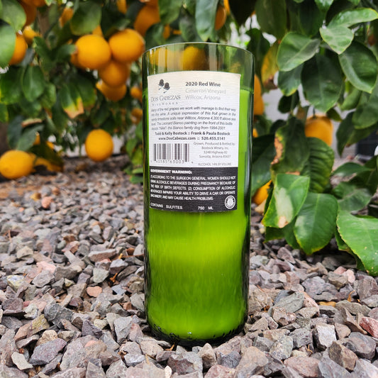Back label of a Dos Cabezas wine bottle candle placed on gravel with a citrus tree in the background. The label and bottle are intact, showcasing the candle's eco-conscious origin and Arizona winery roots.
