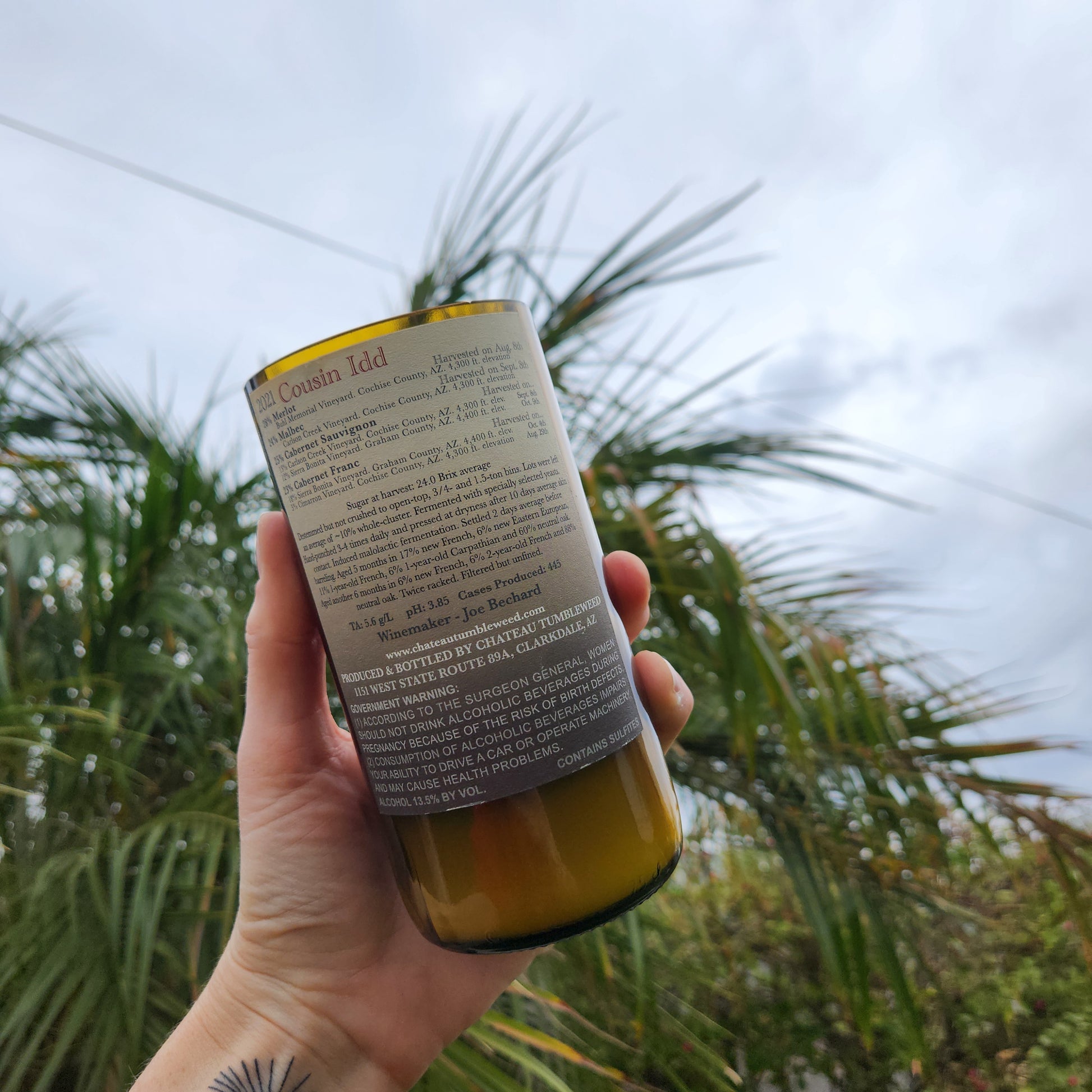 Back label of a recycled wine bottle candle featuring a Chateau Tumbleweed Cousin Idd label. The handmade candle includes a wooden wick and coconut soy wax, captured with Arizona palm trees behind.
