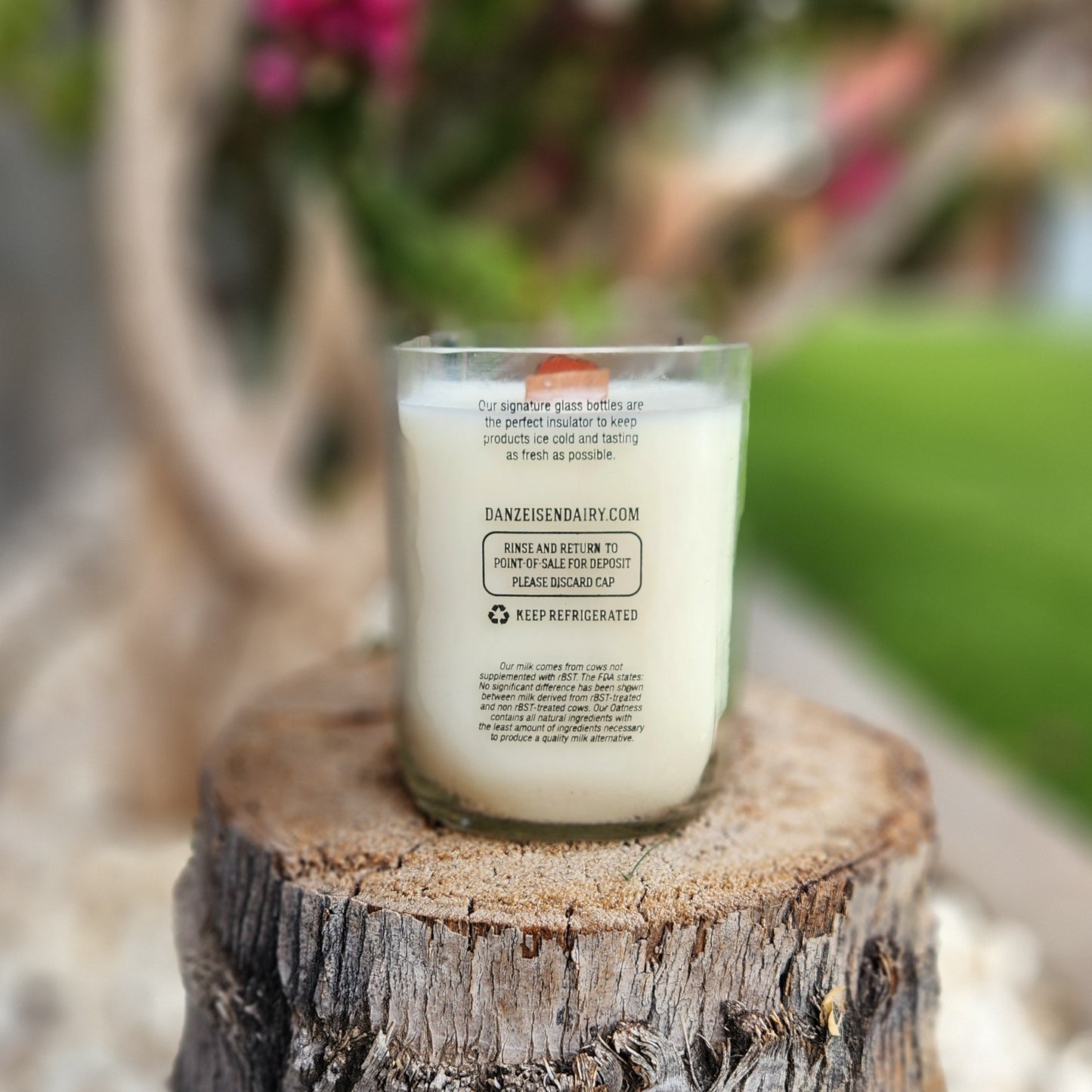 Rear label view of a clean-burning candle made in a repurposed Danzeisen Dairy bottle. Set outdoors on a tree stump, the label details the dairy’s info and highlights the candle’s eco-conscious origin.