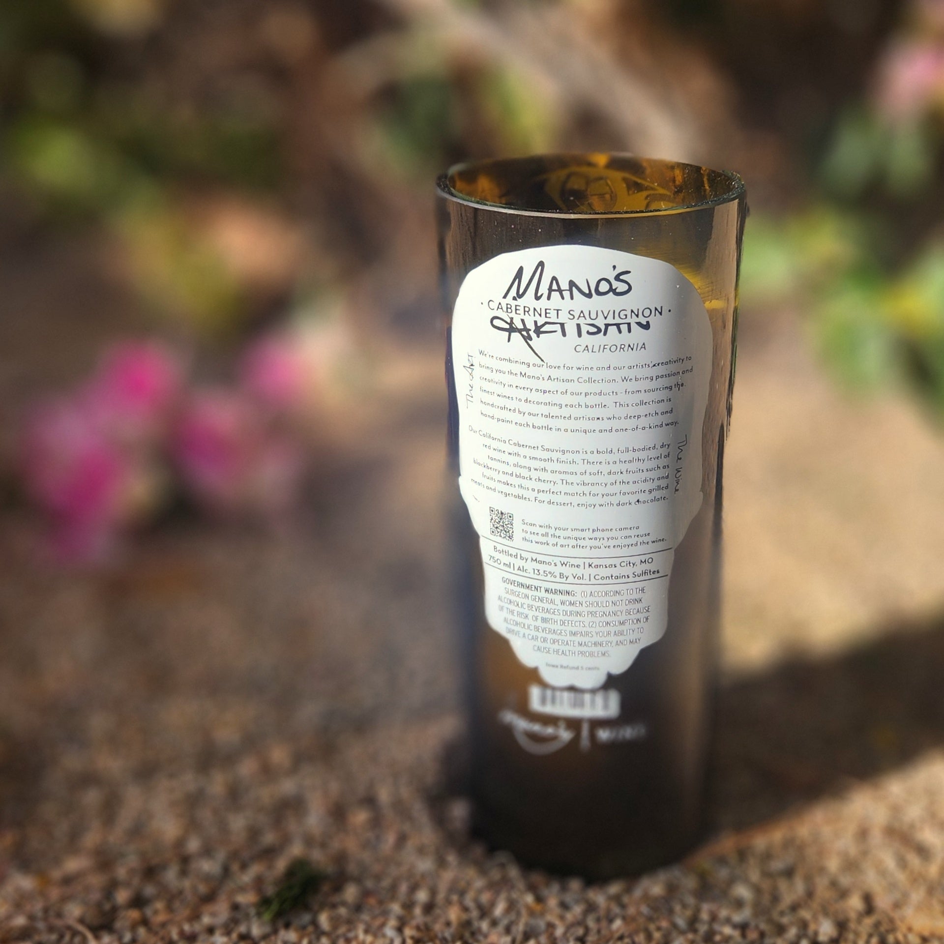 Back label view of a non-toxic, eco-friendly candle poured into a recycled wine bottle, showing a signature from Manos Wine and subtle label wear that adds to its handmade charm.

