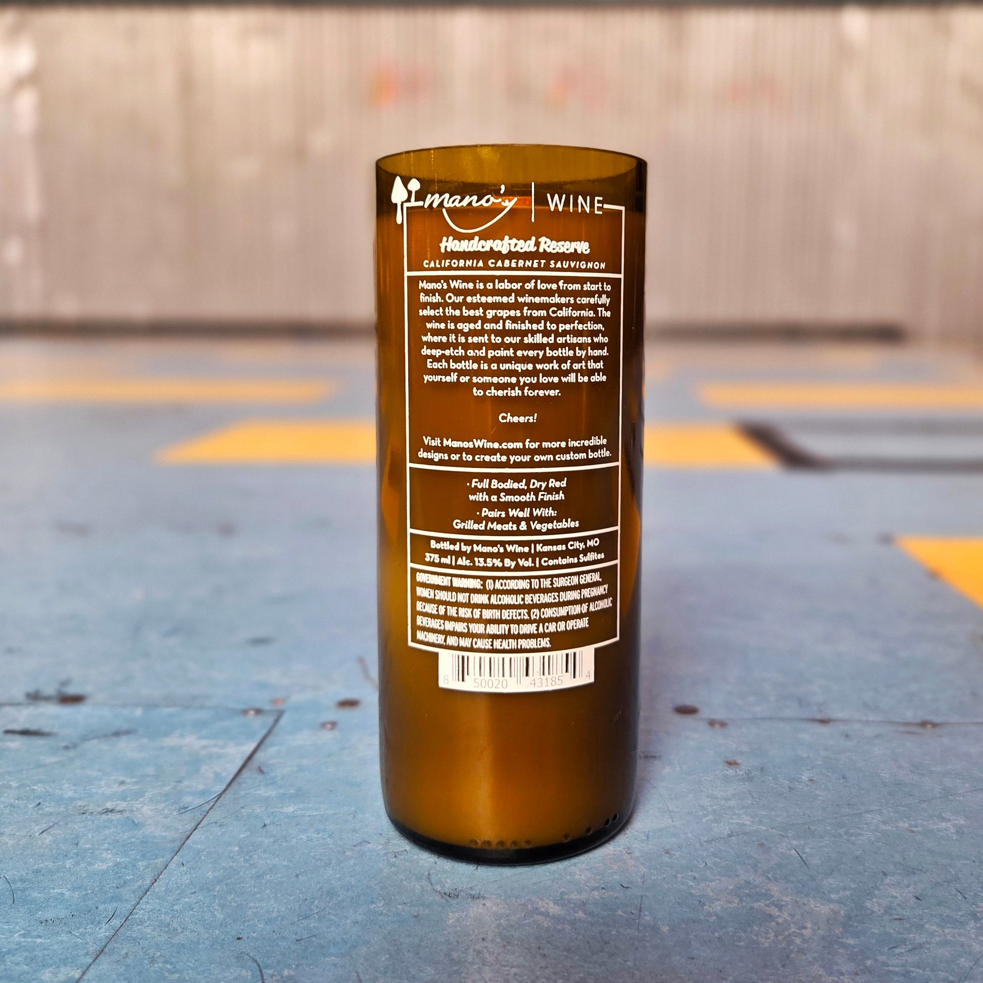 Back view of a recycled wine bottle turned candle showing the original Mano’s Wine label. This upcycled glass now houses a clean-burning, hand-poured coconut soy candle with a wooden wick.