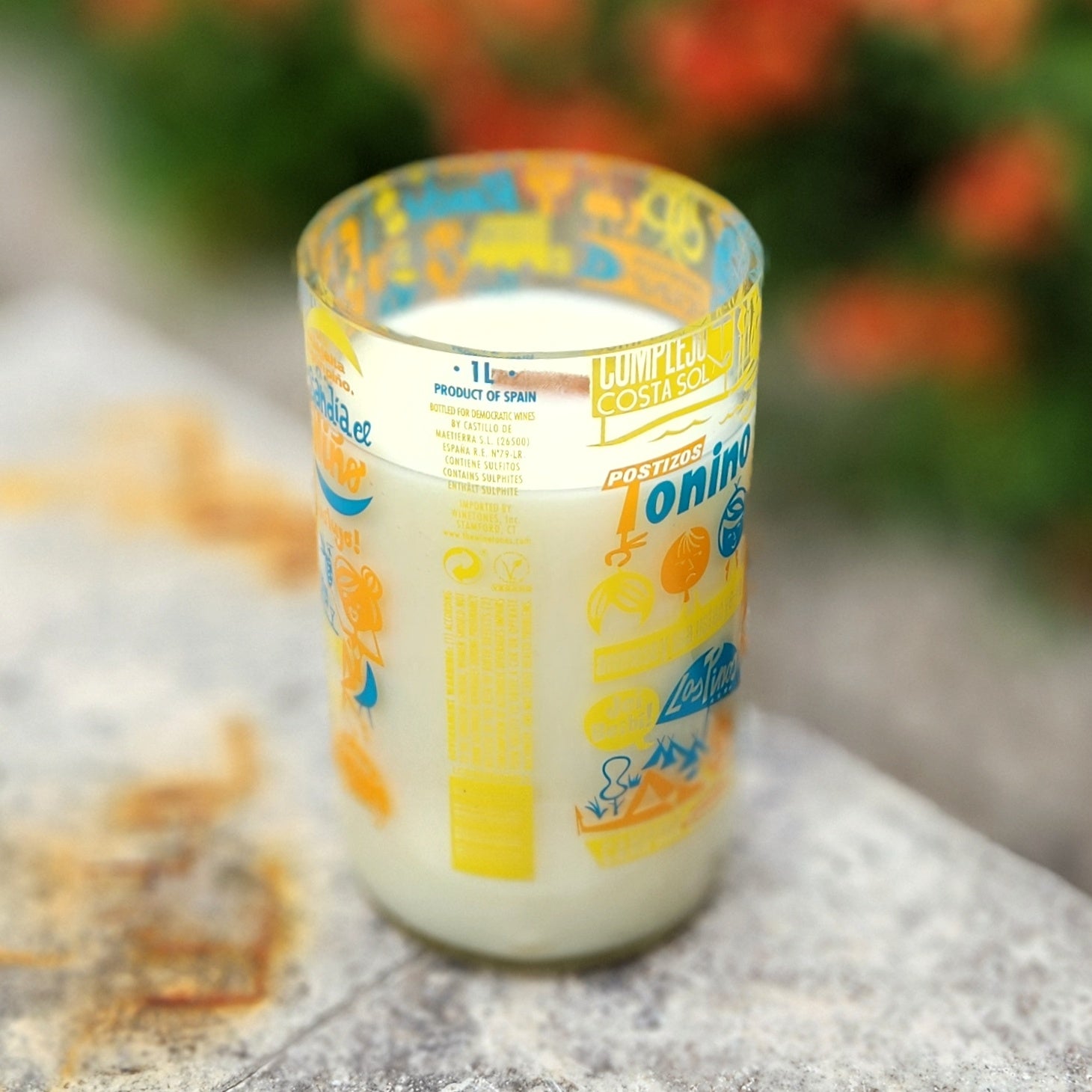 A hand-poured coconut soy wax candle in a recycled wine bottle with a vibrant retro-style travel label in yellow, orange, and blue, featuring whimsical graphics and playful fonts; the candle is placed outdoors on a sunlit stone surface.