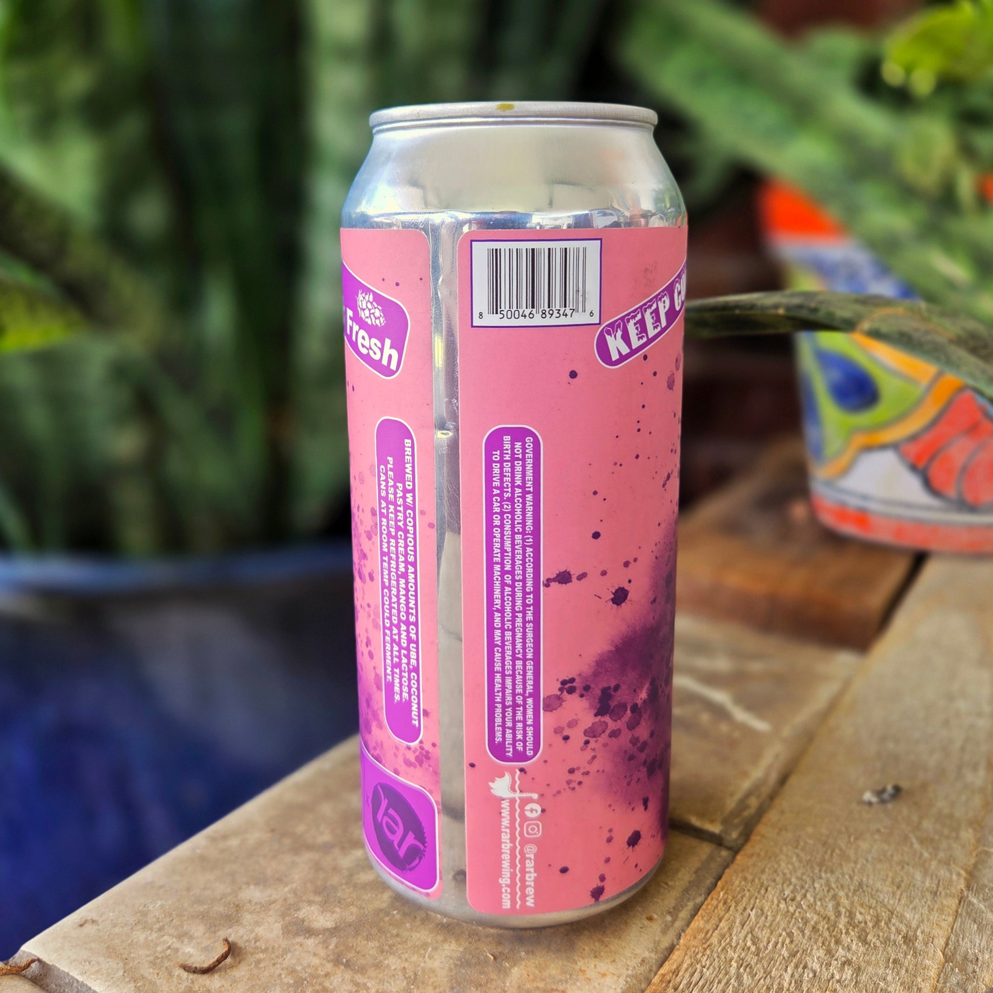 Back side of a recycled beer can candle featuring pink and purple splatter artwork with product details and branding. The can is part of the "Out of BRAAAAAAAAins" zombie dog series and has been upcycled into a coconut soy candle with a wooden wick.