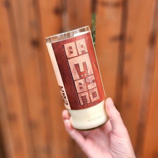 A tilted side view of the bottle showcasing the modern, stacked Bambino text label in earthy tones. The recycled champagne bottle glass is lightly frosted, and the hand holding it is set against a wood backdrop, highlighting its artisan charm.