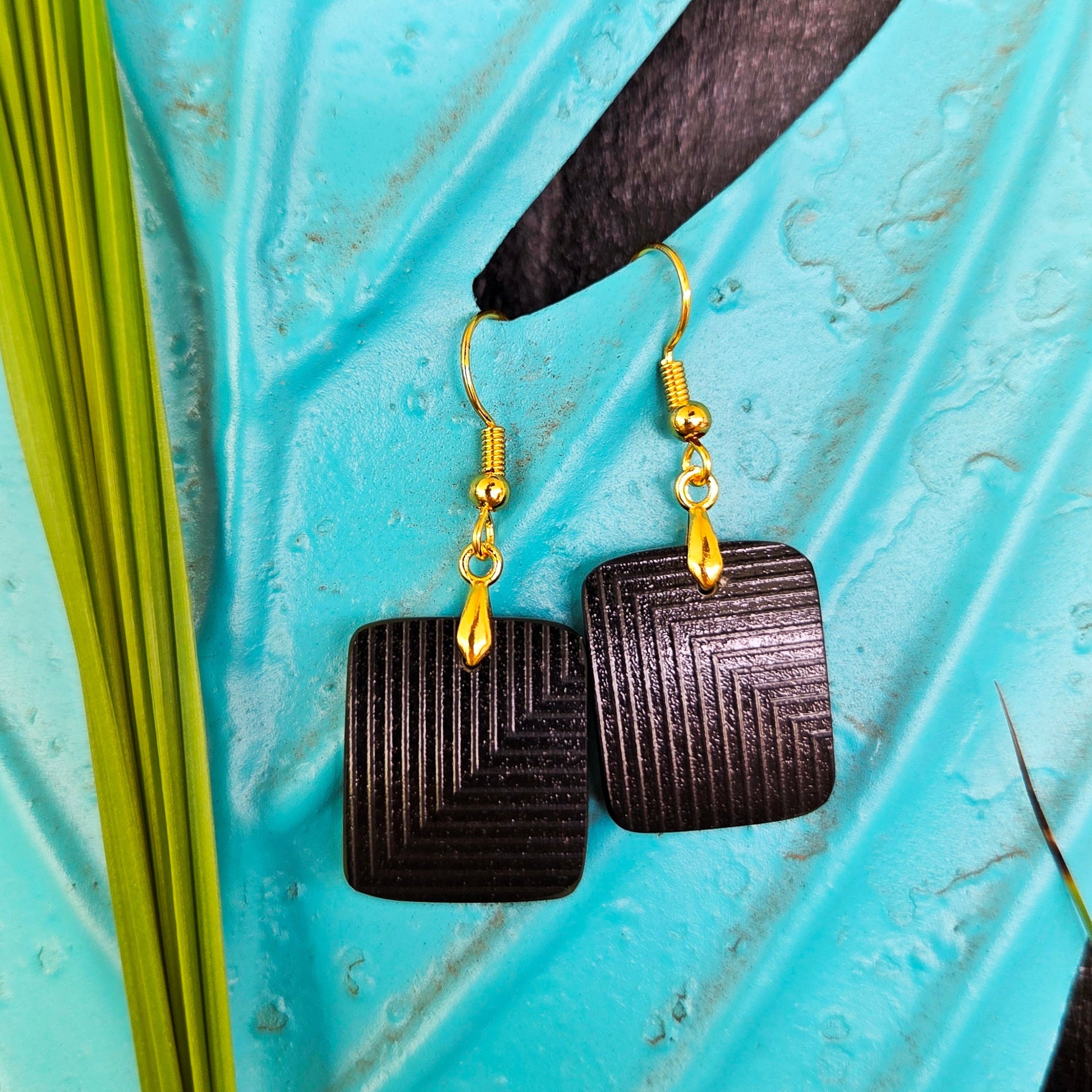 A pair of square black geometric eearrings with gold fishhook wires, displayed on a textured turquoise surface with green leaf.