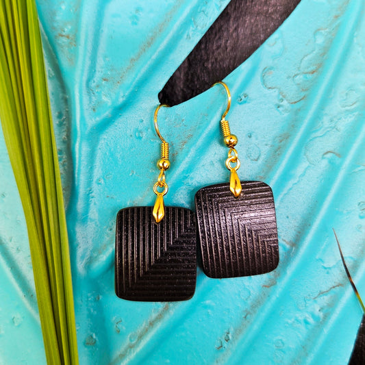 A pair of square black geometric eearrings with gold fishhook wires, displayed on a textured turquoise surface with green leaf.