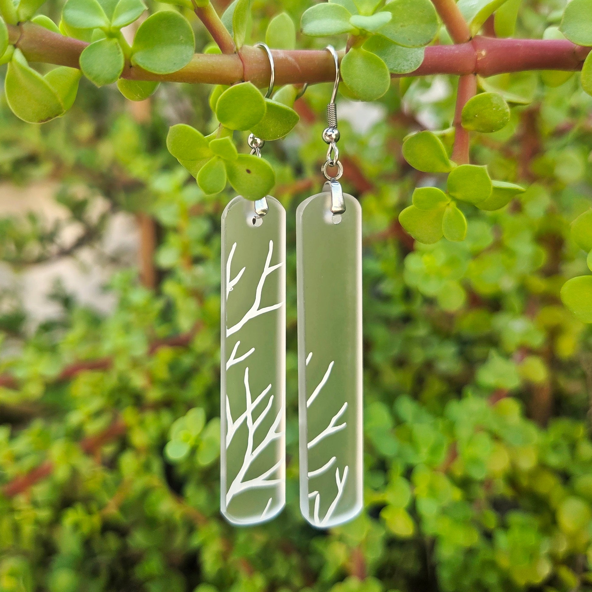 A pair of silver-toned drop earrings with elongated rectangular translucent panels featuring white branch-like line patterns, hanging from a plant branch with green leaves in a natural outdoor setting.