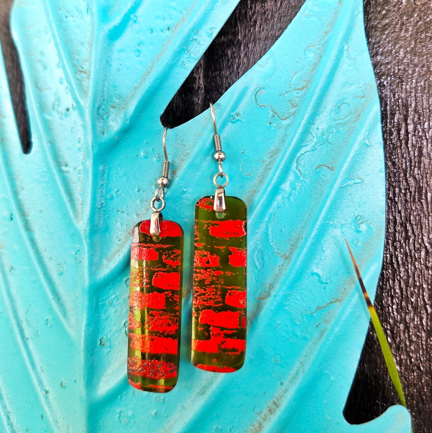 A pair of rectangular drop earrings with a red brick pattern over a green and amber glass background, hanging on silver fishhook wires against a turquoise decorative leaf background.