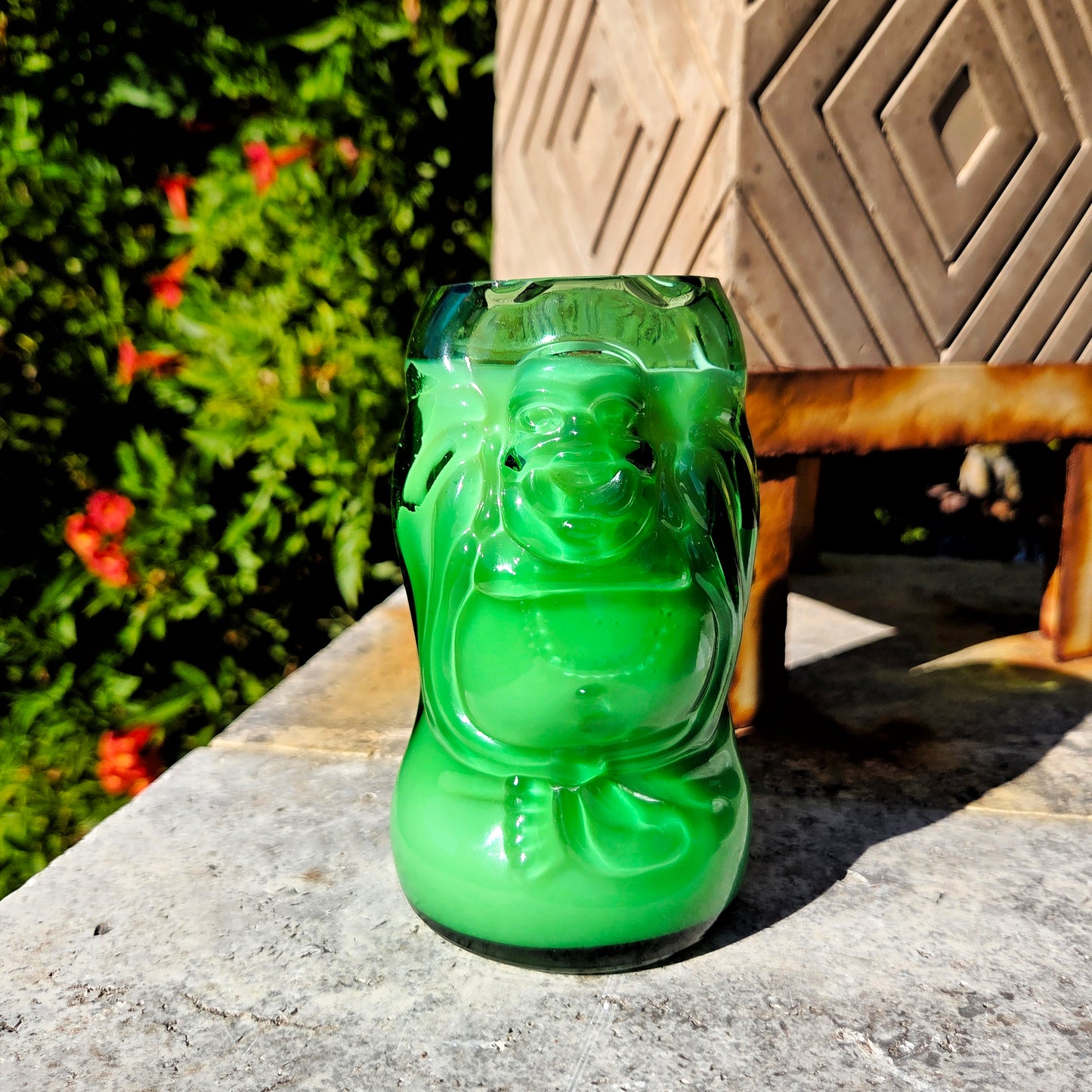 Green glass cut bottle in the shape of a Buddha, make into a candle.  placed on a stone surface in front of greenery and a zen planter.
