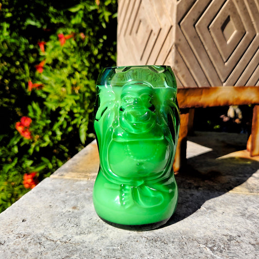 Green glass cut bottle in the shape of a Buddha, make into a candle.  placed on a stone surface in front of greenery and a zen planter.