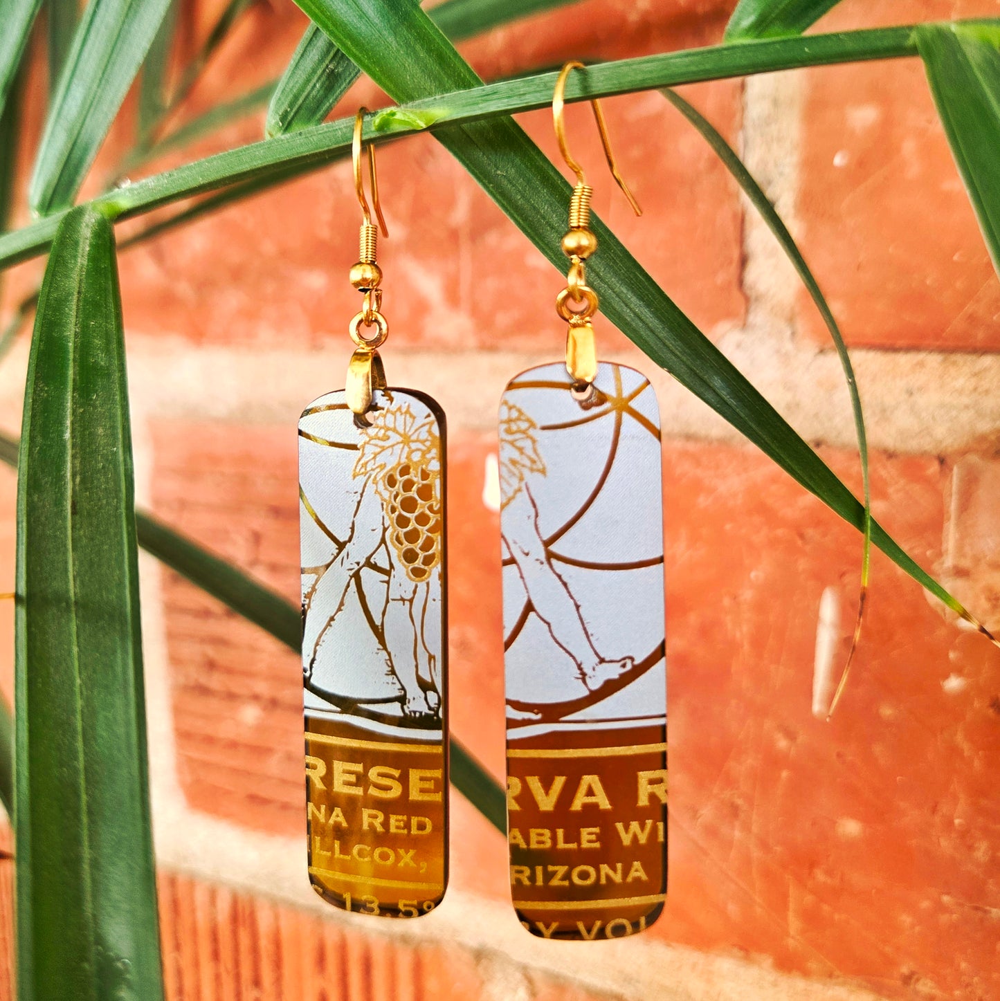 A pair of rectangular drop earrings with gold-tone hooks and gold-tone lettering, featuring a white and gold graphic and wine bottle label design, hanging against a background of green palm leaves and a red brick wall.