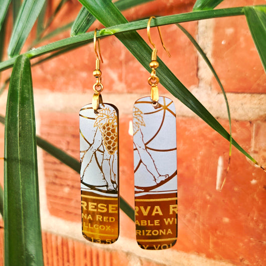 A pair of rectangular drop earrings with gold-tone hooks and gold-tone lettering, featuring a white and gold graphic and wine bottle label design, hanging against a background of green palm leaves and a red brick wall.