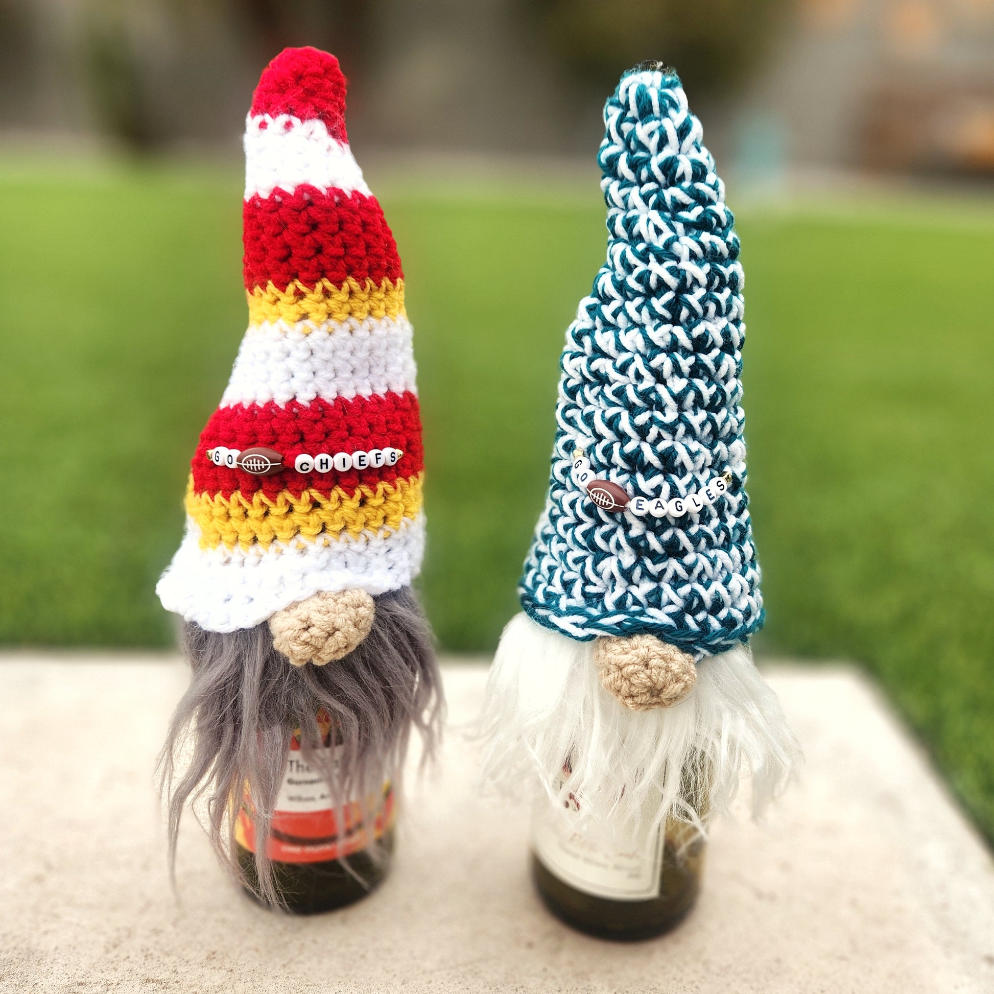 Two wine bottles dressed as whimsical gnomes, each wearing a crochet hat representing football teams. The left gnome sports a red, white, and yellow striped hat with beads spelling "Go Chiefs," while the right gnome wears a green and white hat with beads spelling "Go Eagles." Both have long yarn beards and are set outdoors on a concrete surface with a lush green lawn in the background, creating a playful and sporty atmosphere.