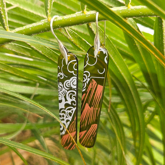 Pair of reclaimed glass earrings with intricate cloud/mountain patterns hanging on green leaves