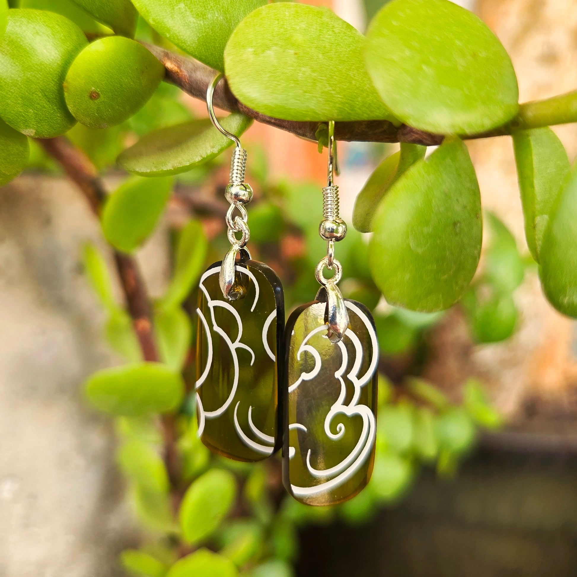 A pair of dangling earrings with rectangular dark green glass pendants featuring white cloud patterns, hanging from silver fishhook wires against a background of green jade plant leaves.