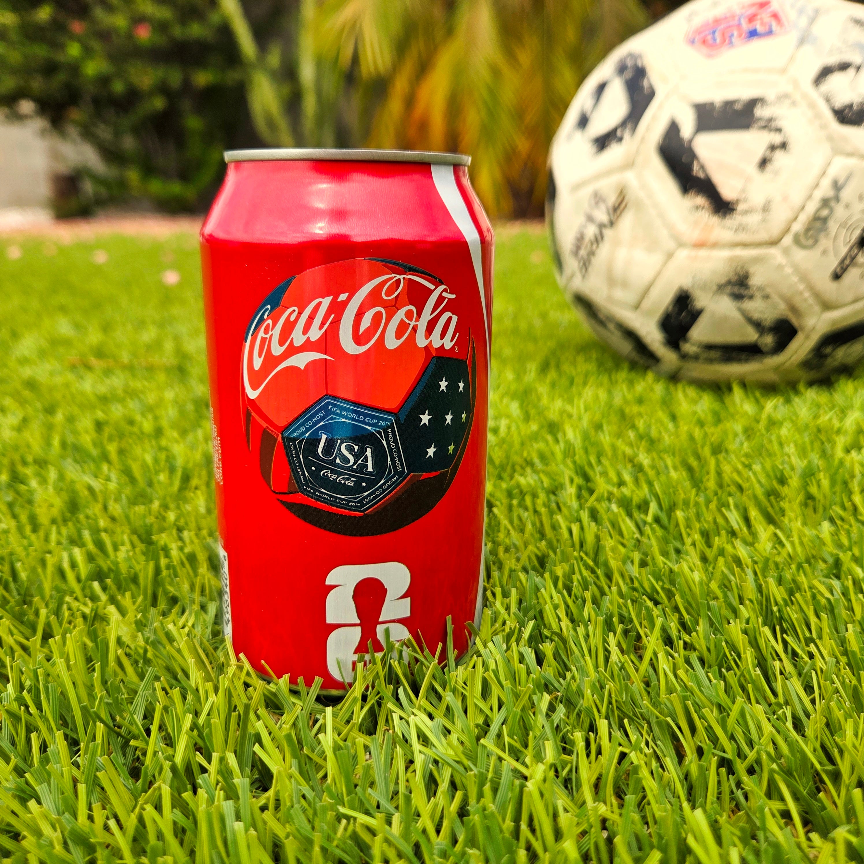 Coke can with USA soccer ball logo sitting on the grass in front of a well used soccer ball.