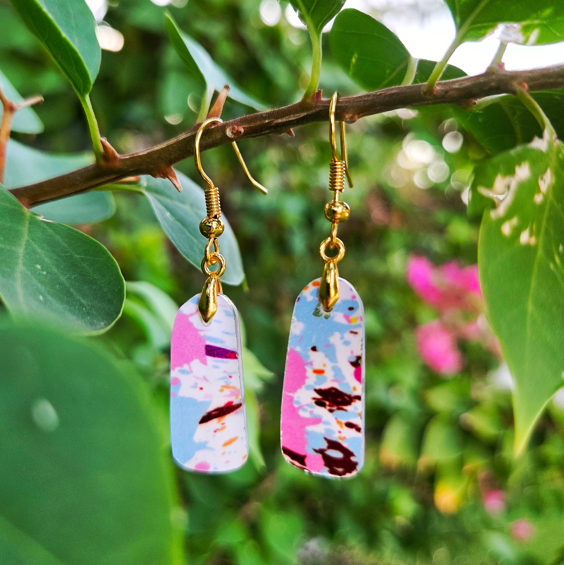 A pair of dangling earrings with gold-tone fishhook wires and rectangular marbled charms in pink, blue, white, and dark brown, hanging from a tree branch with green leaves in the background.