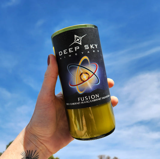 A hand-poured coconut soy wine bottle candle housed in a recycled Deep Sky Vineyards Fusion bottle. The label features a striking atomic fusion design, glowing against a cosmic backdrop. This eco-friendly wooden wick candle has a polished glass rim, showcasing expert craftsmanship. Held up against a vibrant blue sky, the image highlights the candle’s sustainable design and celestial aesthetic, making it a perfect gift for wine lovers and eco-conscious shoppers.