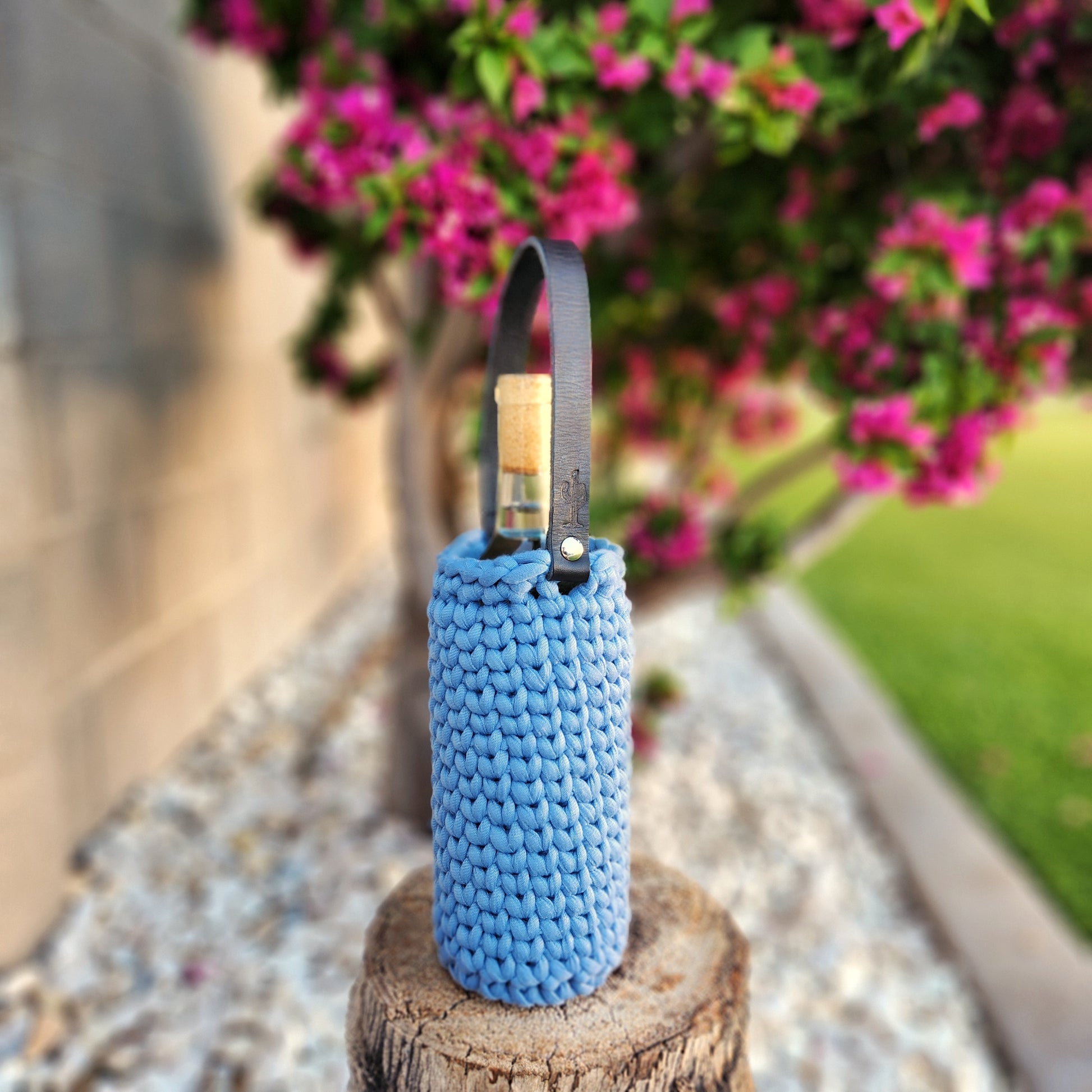 An upcycled wine bottle candle housed in a soft denim blue crocheted carrier with a sturdy black leather handle, also stamped with a cactus symbol. The candle is positioned on a tree stump with pink bougainvillea and greenery in the background, creating a bright and inviting outdoor scene.