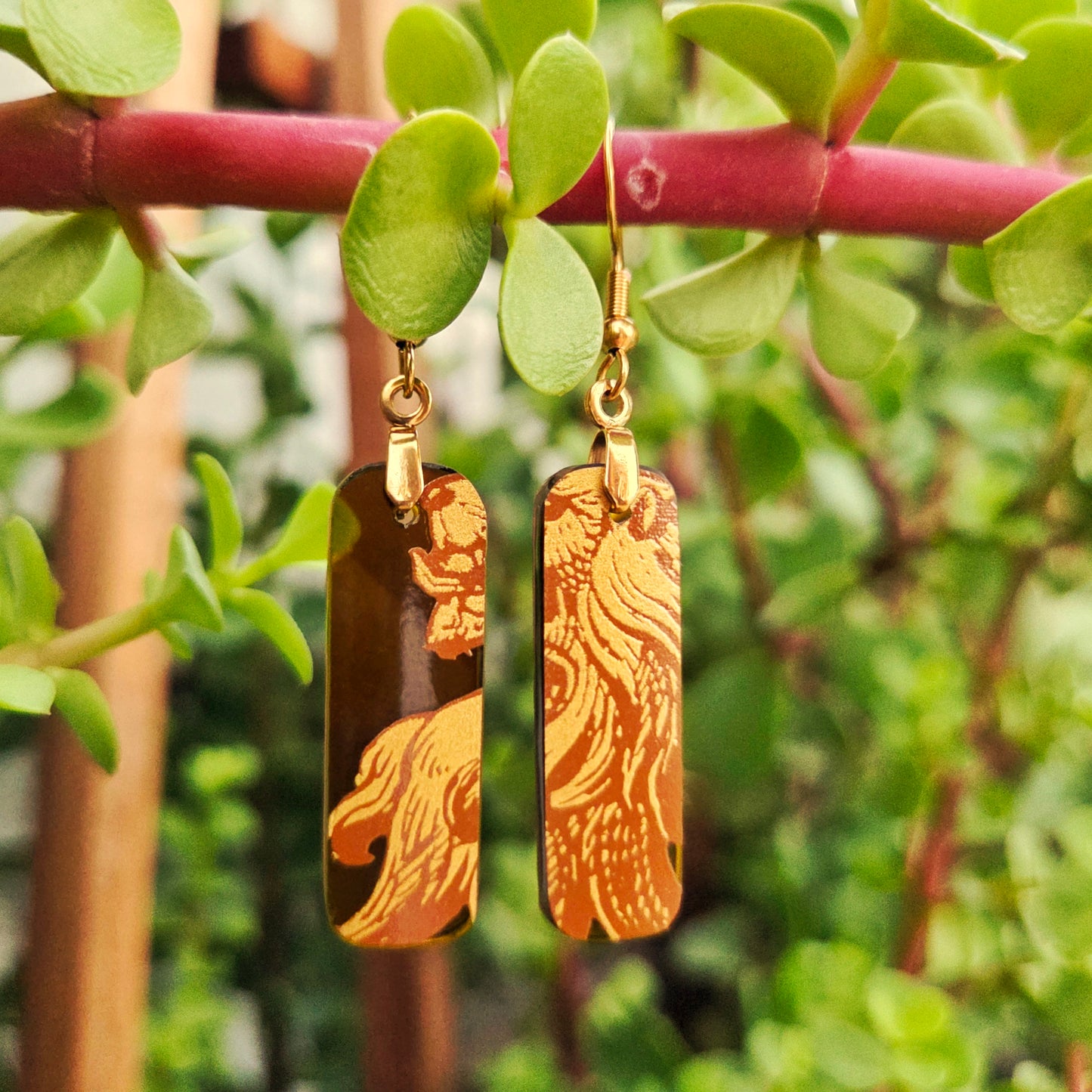 A pair of rectangular drop earrings with gold-toned lion engravings on a dark brown background, hanging from gold hooks and suspended in front of green leaves and a reddish stem.
