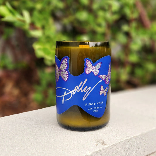Front view of a sustainable candle in a green wine bottle featuring a bright blue label with Dolly Parton's name and purple butterflies, highlighting the upcycled nature of the glass and the non-toxic wax formula