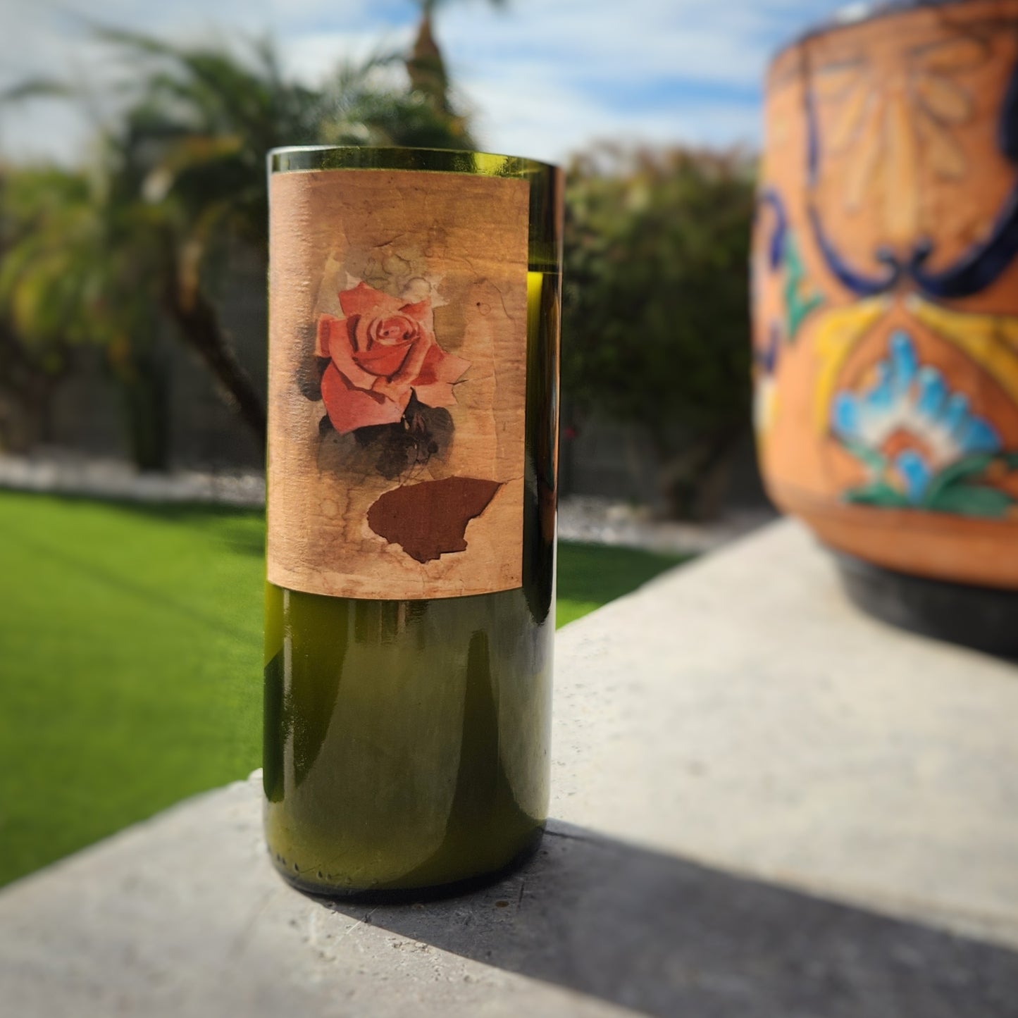 Highlighting timeless elegance, this hand-poured candle in a recycled wine bottle features a rustic rose design. Set against a sunny outdoor scene with vibrant greenery and colorful pottery, it embodies sustainability and artistry