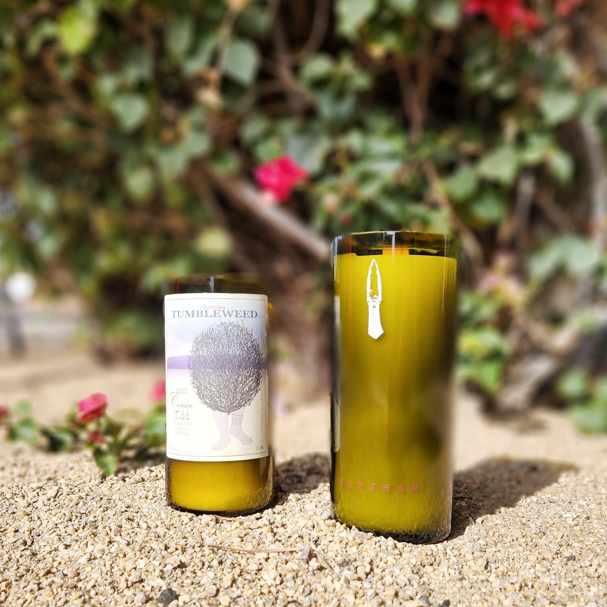 Eco-friendly candles hand-poured into recycled wine bottles, featuring wooden wicks and distinct labels—one from Chateau Tumbleweed and the other  a minimalist bottle. Clean-burning and sustainable, set against a sunny desert garden backdrop.