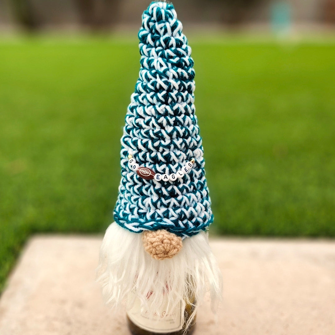 A decorative gnome with a tall crocheted te aland white hat, white beard, and beige nose, standing on a stone surface with a blurred green lawn background. Beads on the hat say "Go Eagles" with a football charm.