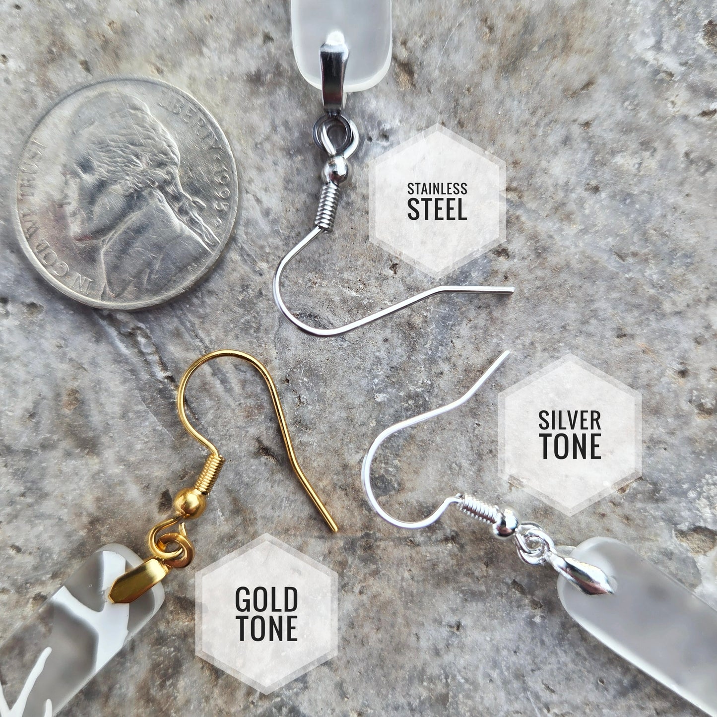 Four types of dangle earrings displayed on a textured stone surface next to a US quarter coin for scale. The earrings include: a stainless steel wire hook with a clear resin drop, a gold-tone fishhook with a clear resin drop, a silver-tone fishhook with a clear resin drop, and a silver-tone fishhook with a faceted frosted white glass drop.