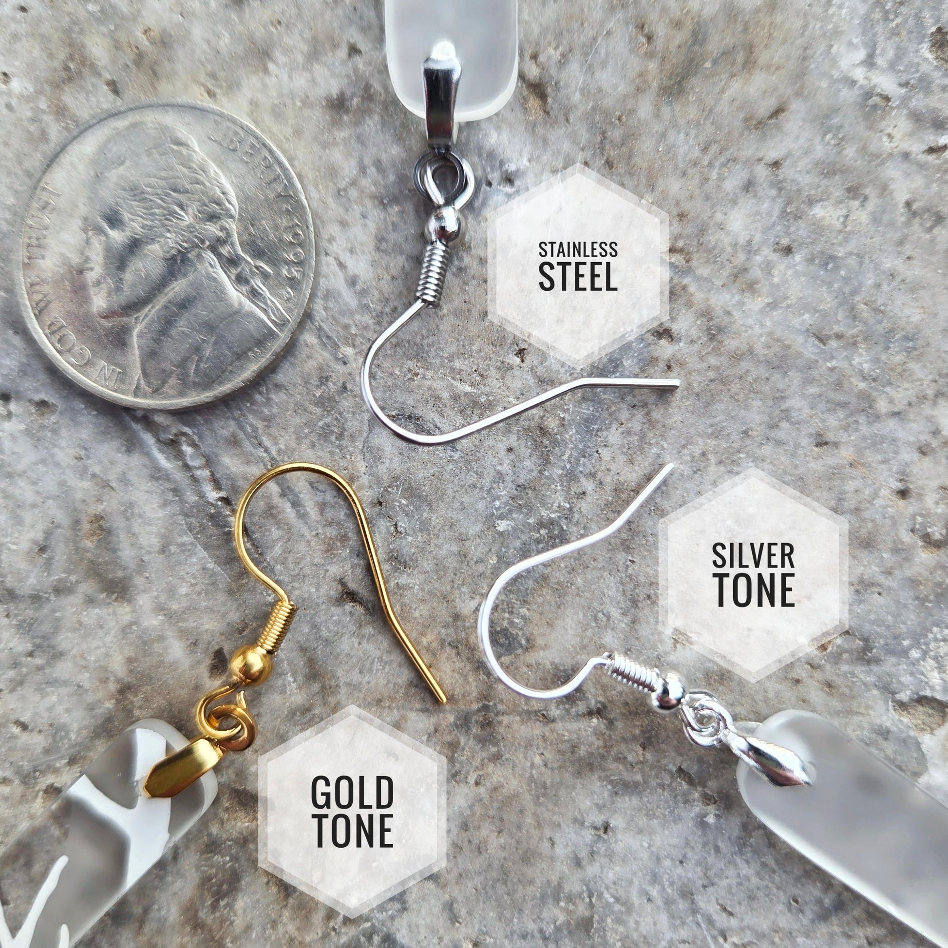 Four types of dangle earrings displayed on a textured stone surface next to a US quarter coin for scale. The earrings include: a stainless steel wire hook with a clear resin drop, a gold-tone fishhook with a clear resin drop, a silver-tone fishhook with a clear resin drop, and a silver-tone fishhook with a faceted frosted white glass drop.