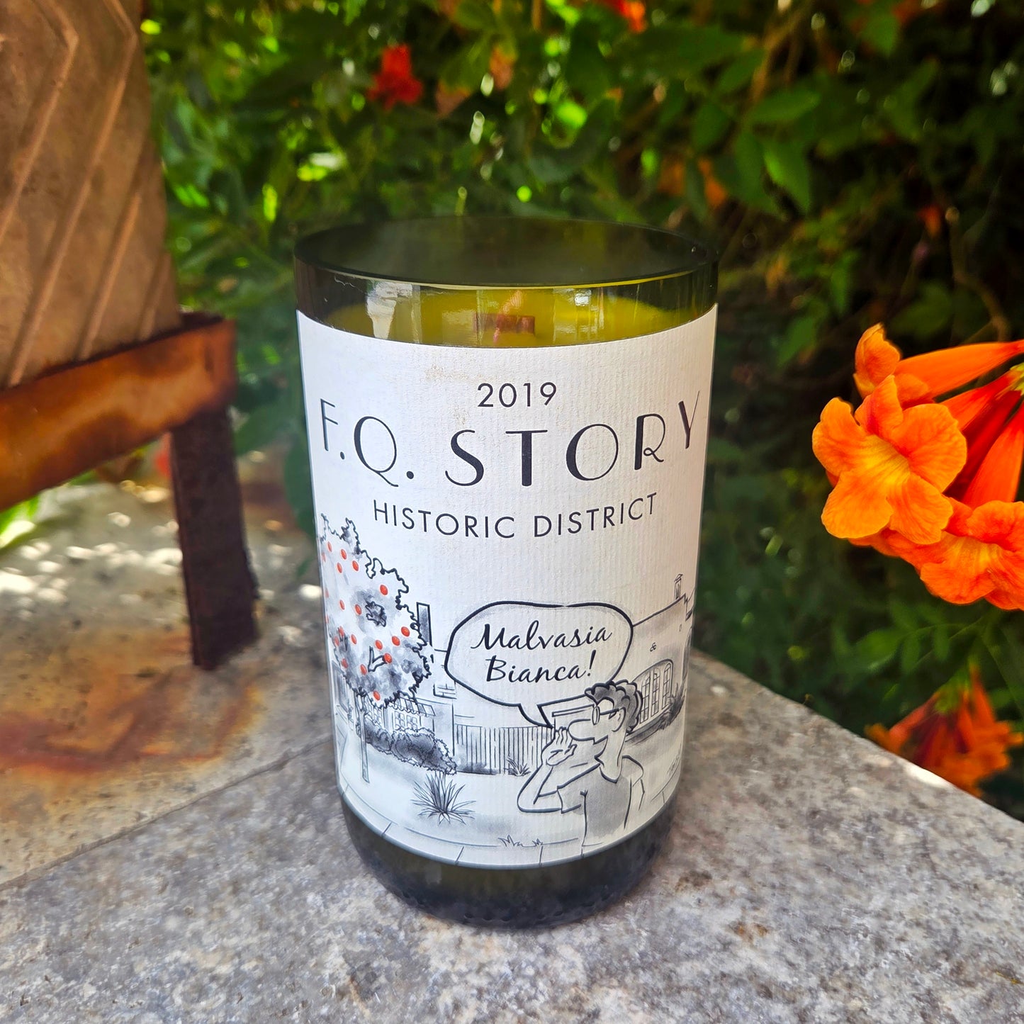 Wine bottle candle featuring artwork of house and orange tree with a cartoon saying "malvasia bianca".  Candle is labeled FQ Story Historic District and is sitting outdoors.