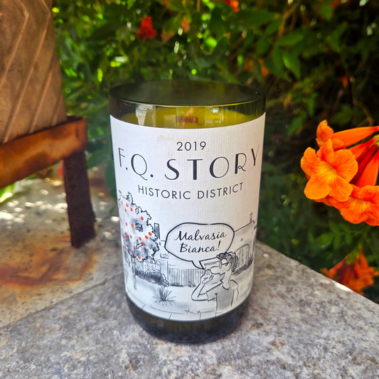 Wine bottle candle featuring artwork of house and orange tree with a cartoon saying "malvasia bianca".  Candle is labeled FQ Story Historic District and is sitting outdoors.