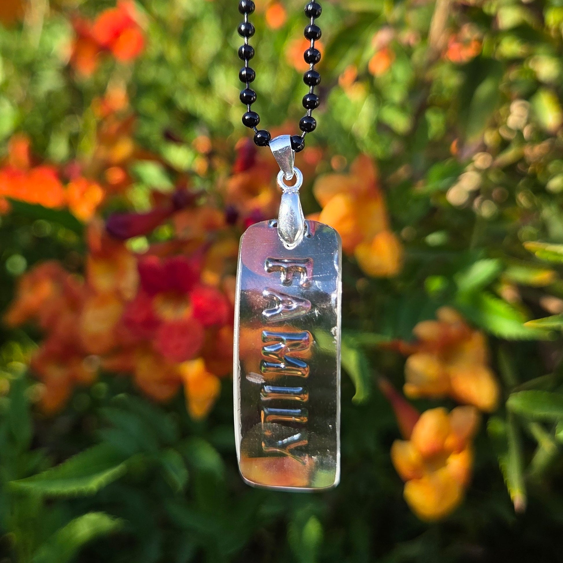 Clear Glass necklace with raised letters spellingt family.  Hanging on a black ball chain in front of orange and green foliage.