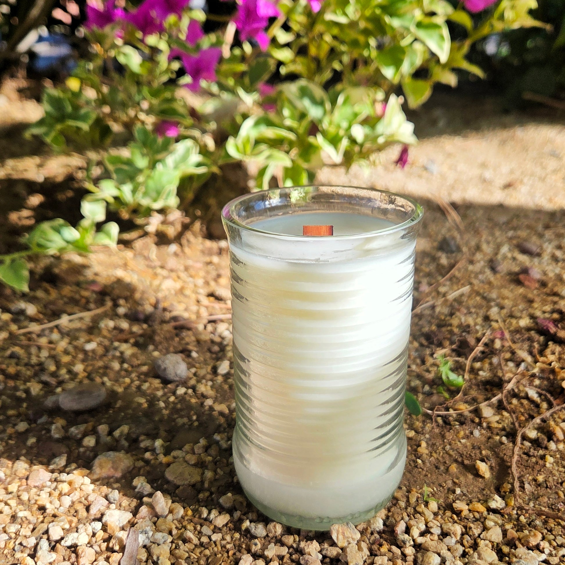 Candle in a glass container on a pebbly surface with plants in the background
