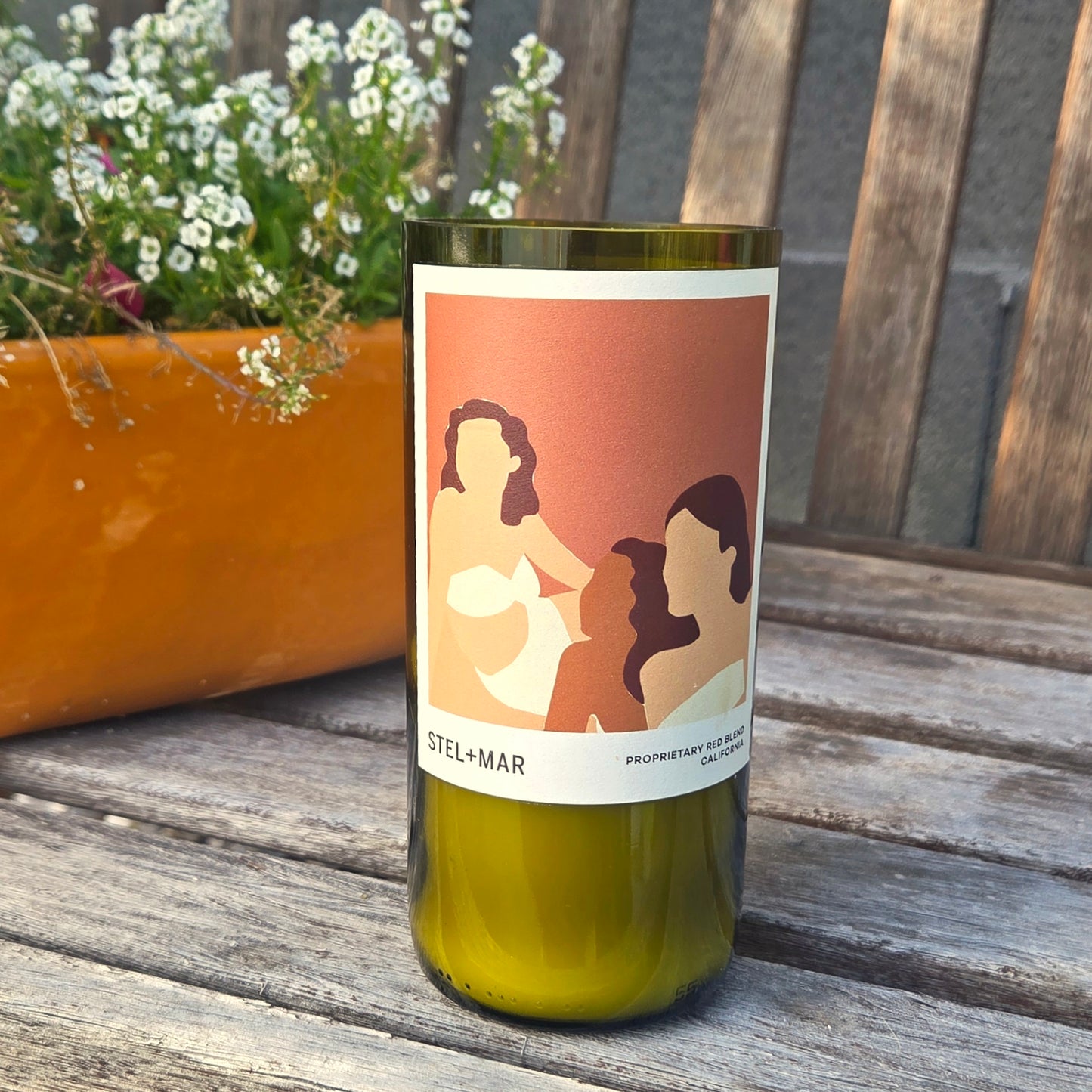 Wine Bottle CAndle with abstract art of women on the label.  Candle is on a weathered wooden bench with flowers in the background.