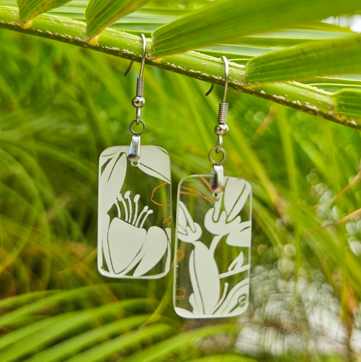 A pair of rectangular dangling earrings with a white floral cutout design, hanging from silver fishhook wires against a blurred green tropical foliage background.