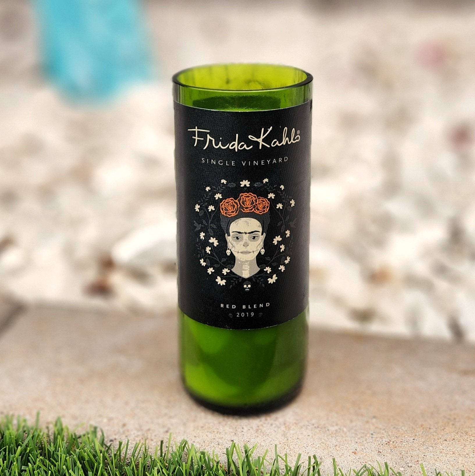 Non-toxic candle in a recycled wine bottle featuring Frida Kahlo’s portrait with floral crown and white flowers on a matte black label, filled with coconut soy wax and topped with a crackling wooden wick.

