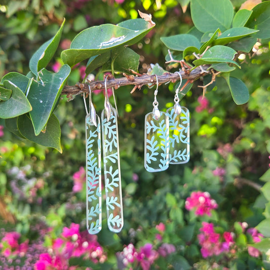 Transparent glass earrings made from reclaimed liquor bottles with vine patterns hanging on a branch with green leaves and pink flowers.
