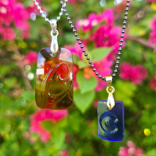 2 glass penants hanging on ball chains featuring the letter C.  One in amber glass, the other in blue, in front of greenery with pink flowers.
