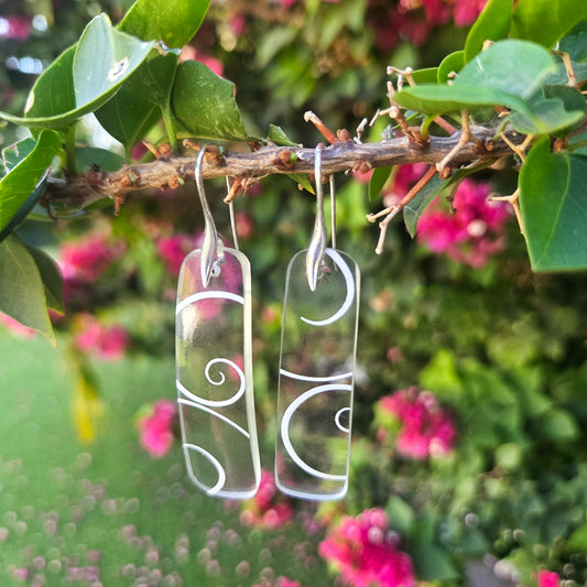 Clear glass wine bottle earrings with white patterns hanging on a branch with green leaves and pink flowers.